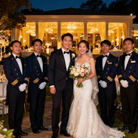 一对新人在婚礼结束后与金狮礼宾团队合影，新人站在中间手捧鲜花笑容灿烂，身后是六位身着统一制服的礼宾专员，背景是夜色中灯火辉煌的宴会厅外景。